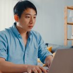 Freelance Asia guy casual wear using laptop online learning in living room at house. Working from home, remotely work, distance education, social distancing, quarantine for corona virus prevention.